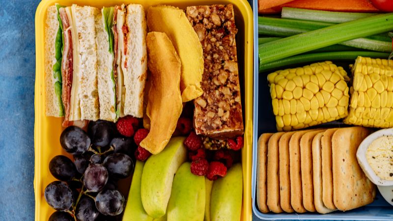 Lancheiras amarela e azul com sanduíches, frutas, verduras e biscoitos.